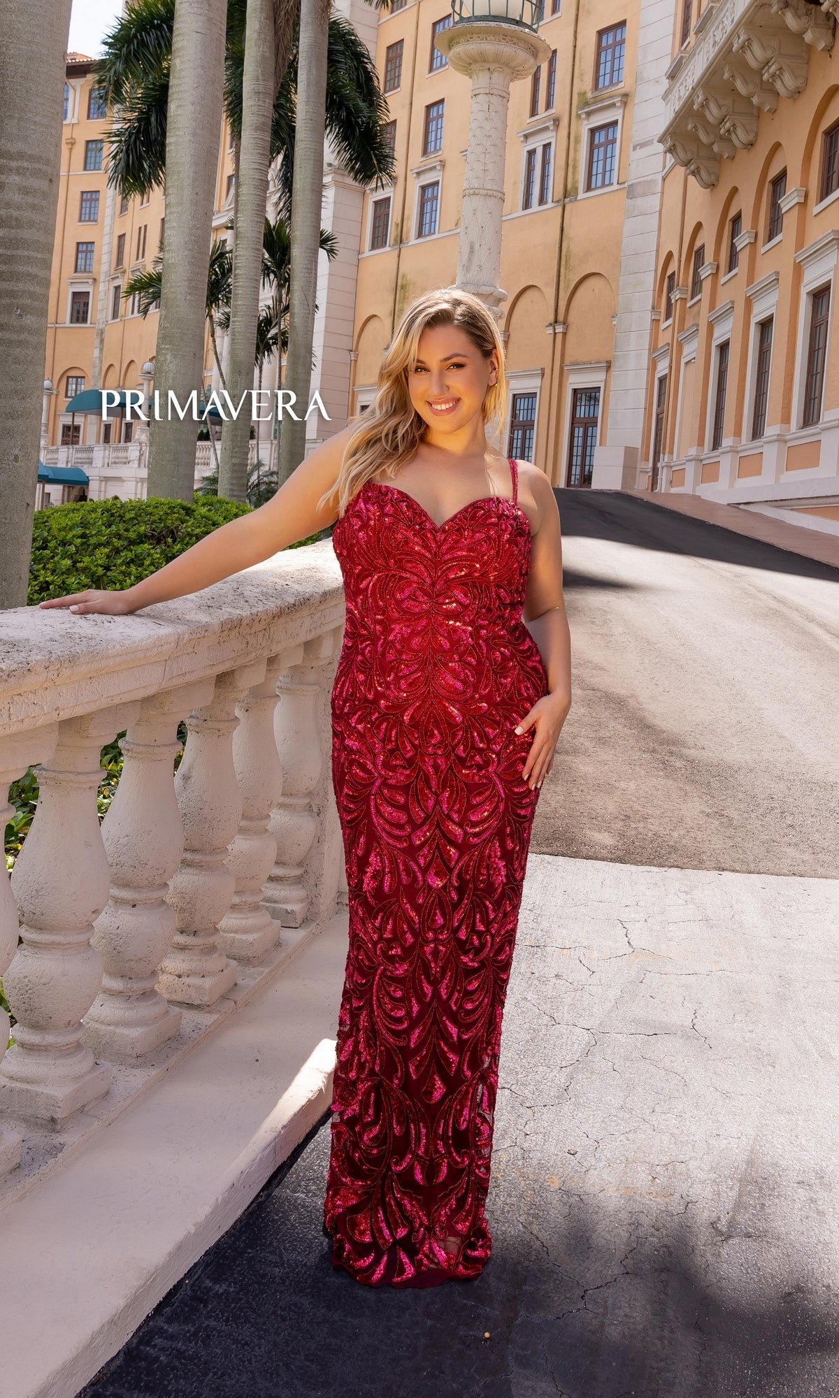 A woman wears the Primavera Long Plus-Size Beaded Prom Dress 14054, standing outdoors by a white stone railing with a beige building and palm trees in the background.